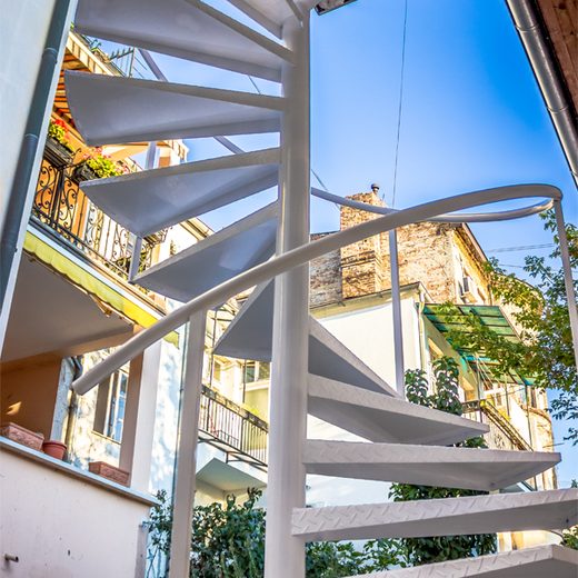 Pogled odozdo na belo spoljno spiralno metalno stepenište sa rebrastim gazištima u gradskom dvorištu